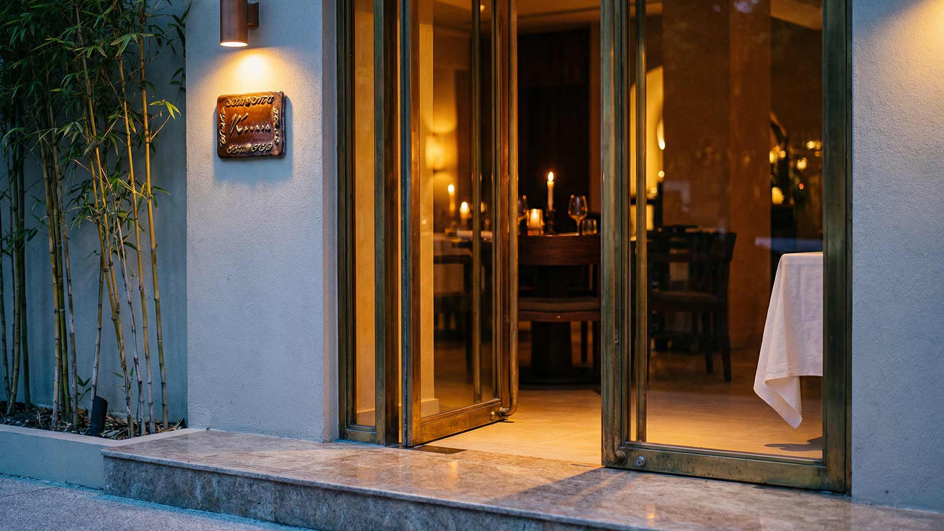 Elegant villa office entrance at dusk in Cherng Talay with brass-framed glass doors, bamboo plantings, and warm candlelit interior
