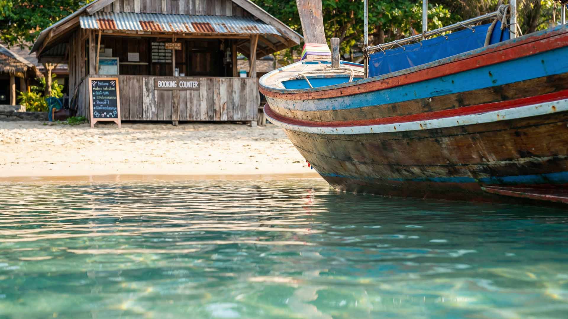 Weathered blue-and-red longtail boat bow in crystal-clear shallows with a rustic wooden booking counter and chalkboard menu on the sandy shore behind at Kata Beach