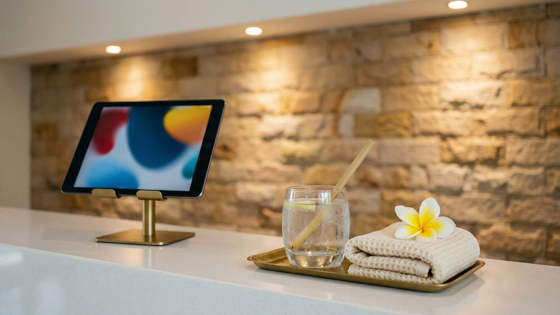 iPad on a brass stand beside a lemongrass-infused welcome drink, folded waffle towel, and frangipani flower on a white quartz spa reception counter with warm sandstone accent wall