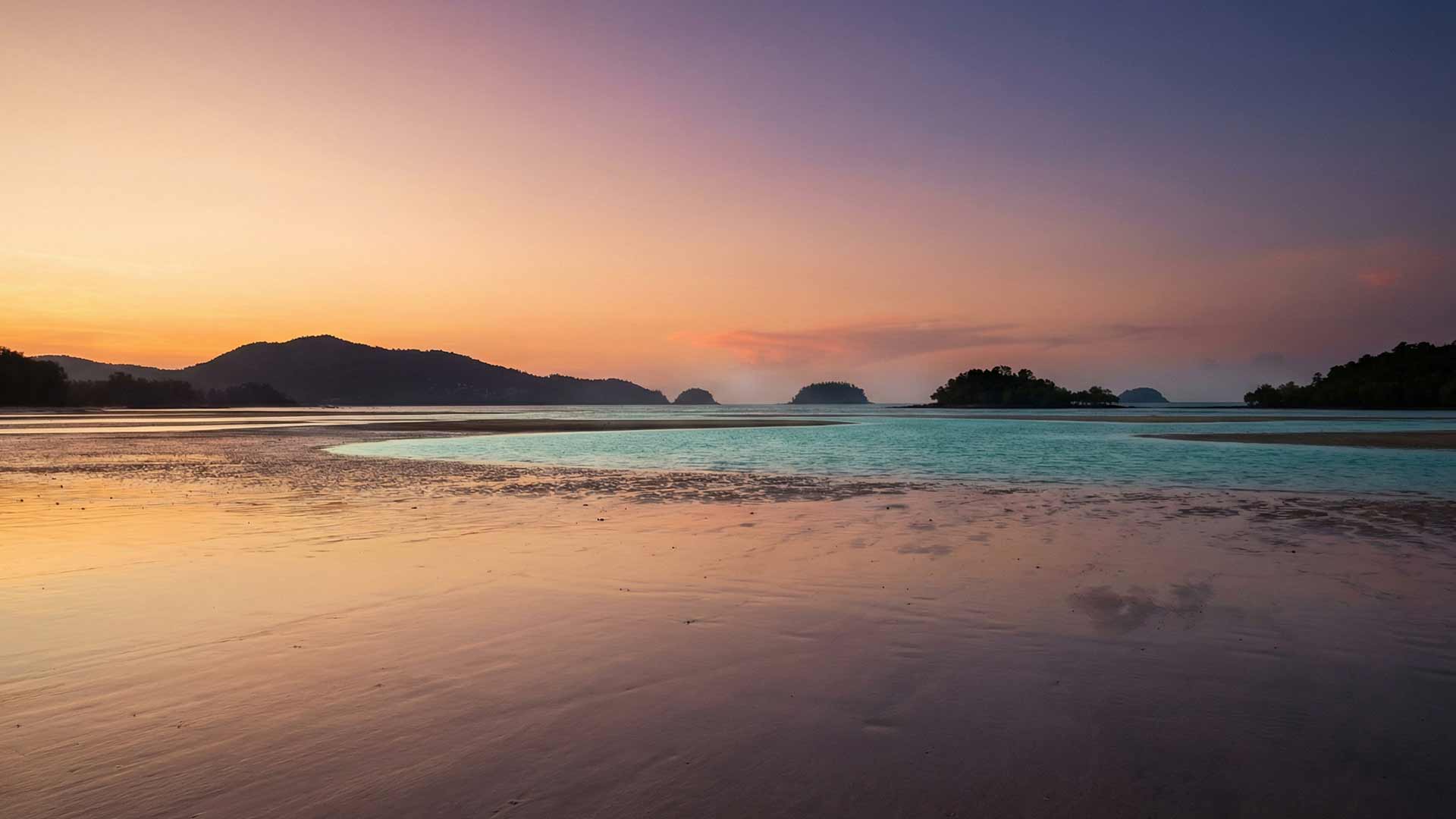 Panoramic sunset over Bang Tao Bay at low tide with turquoise shallows, mountain silhouettes, and warm amber-pink sky reflections
