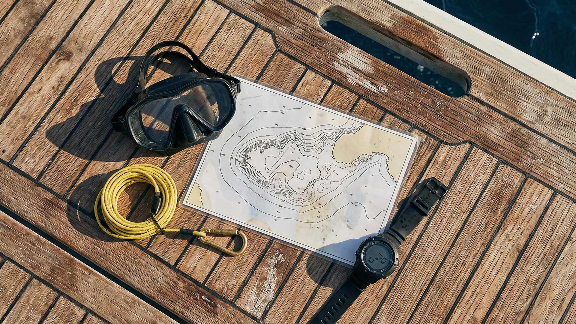 Overhead flat-lay of a dive mask, bathymetric chart, coiled yellow lanyard with brass carabiner, and a dive computer on sun-bleached teak decking beside dark Andaman water at Chalong Bay