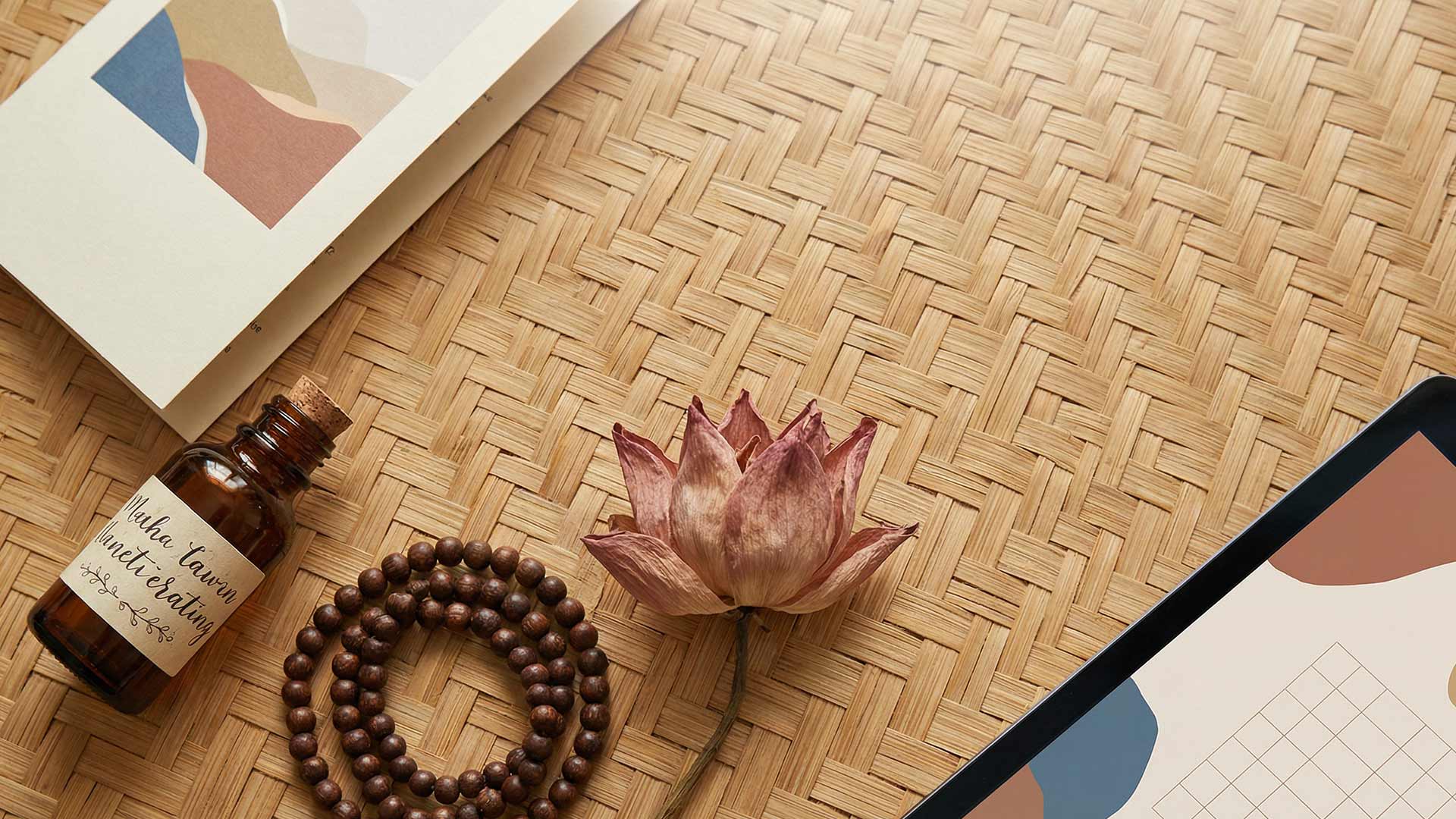 Flat lay on woven bamboo mat with amber essential oil bottle, wooden mala beads, dried lotus flower, minimalist art print, and tablet showing wellness brand wireframes