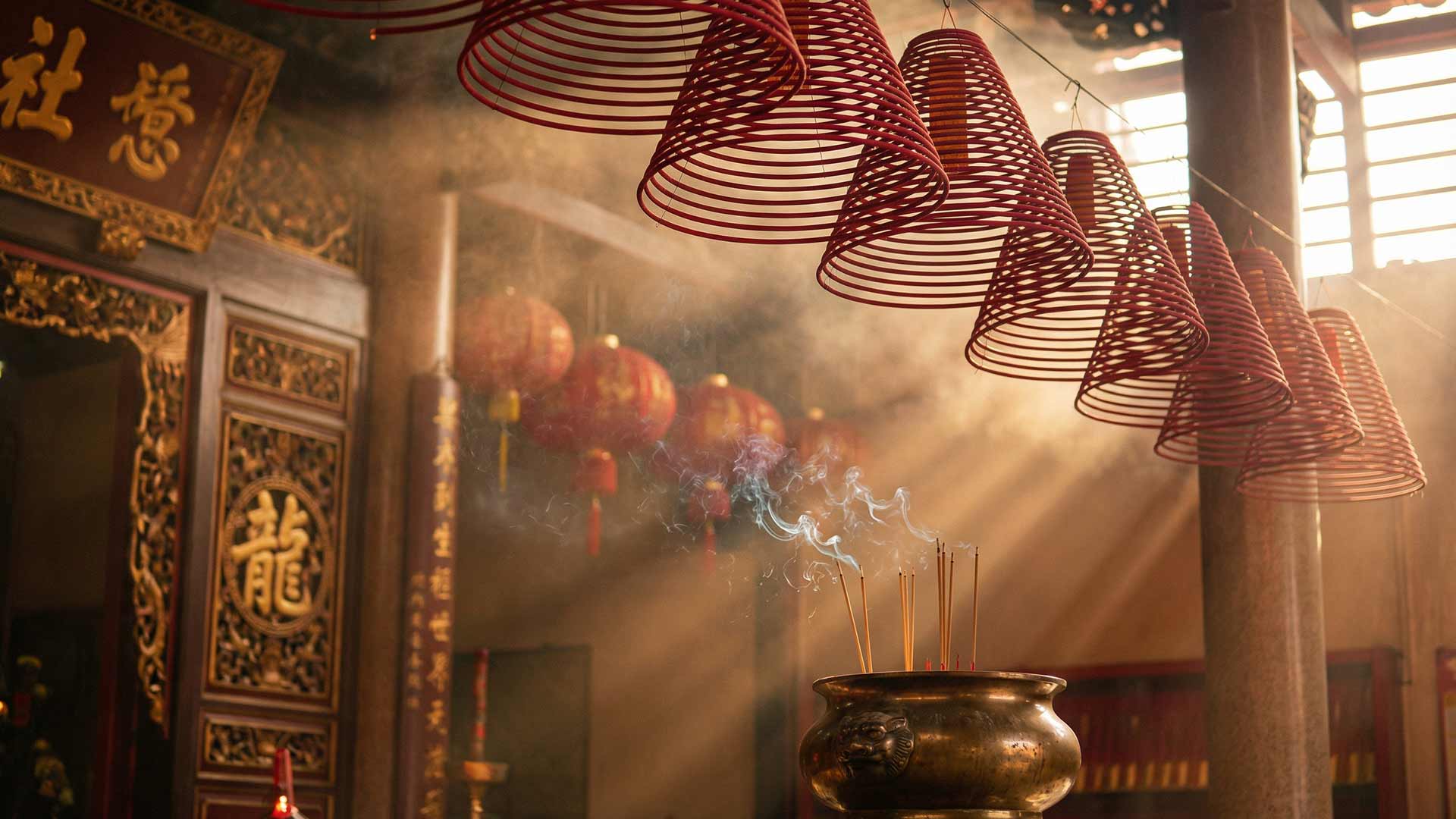 Incense coils and smoke curling through golden light inside a traditional Chinese shrine in Phuket Old Town with red lanterns