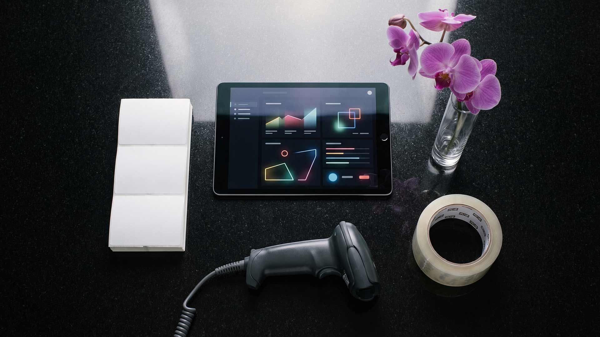 Flat lay on dark granite surface with tablet displaying colorful e-commerce analytics dashboard, barcode scanner, shipping labels, orchid in glass vase, and packing tape