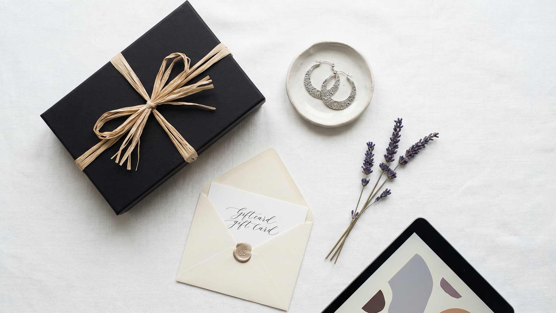 Luxury flat lay with matte-black gift box, wax-sealed gift card envelope, silver earrings, lavender sprigs, and tablet on white linen