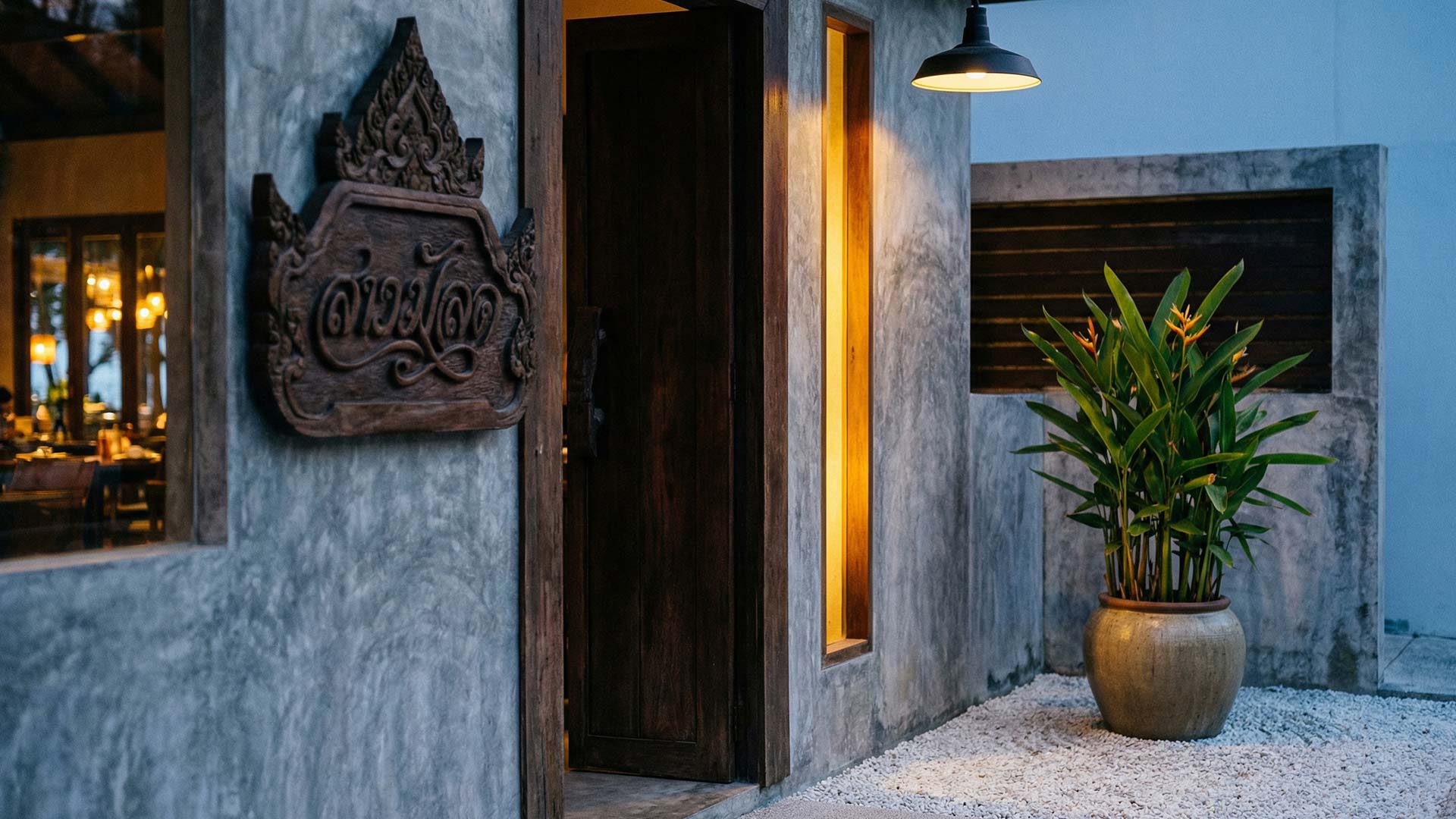 Hand-carved Thai script signboard on a polished cement wall beside a warm-lit teak doorway, industrial pendant lamp, and a ceramic pot of heliconia at a Bang Tao beach club entrance
