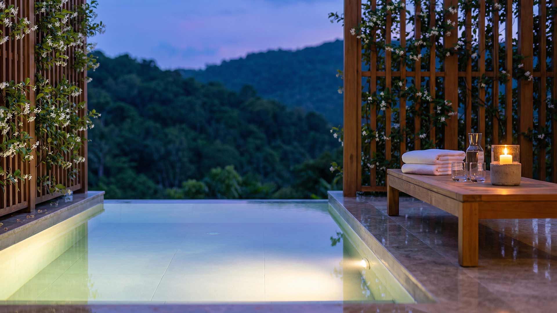 Private infinity plunge pool at a Nai Harn hillside villa at dusk with jasmine-covered timber screens, folded towels, candle, and forested mountains in the background