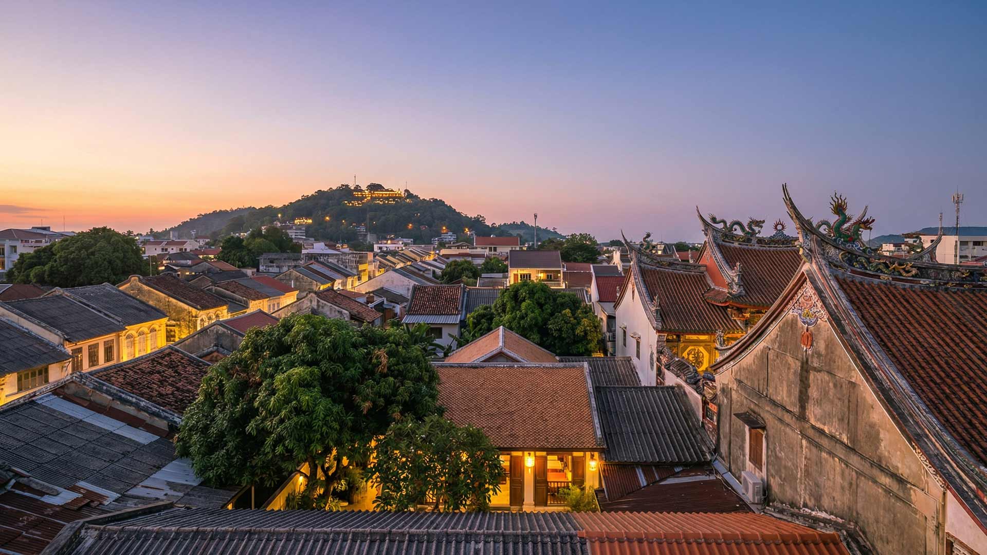 Panoramic rooftop view of Phuket Old Town at dusk with terracotta rooftops, Chinese shrine dragon finials, and Rang Hill glowing on the horizon