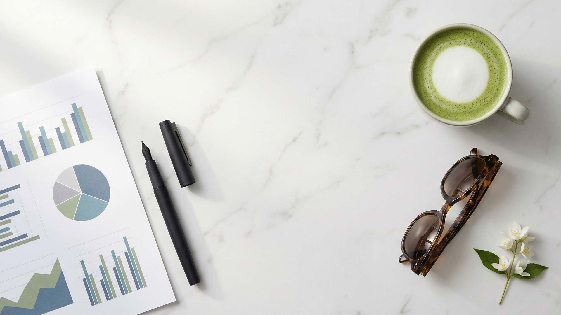 Flat-lay analytics report with bar charts and pie graphs beside a black fountain pen, tortoiseshell sunglasses, matcha latte, and jasmine sprigs on a white marble tabletop