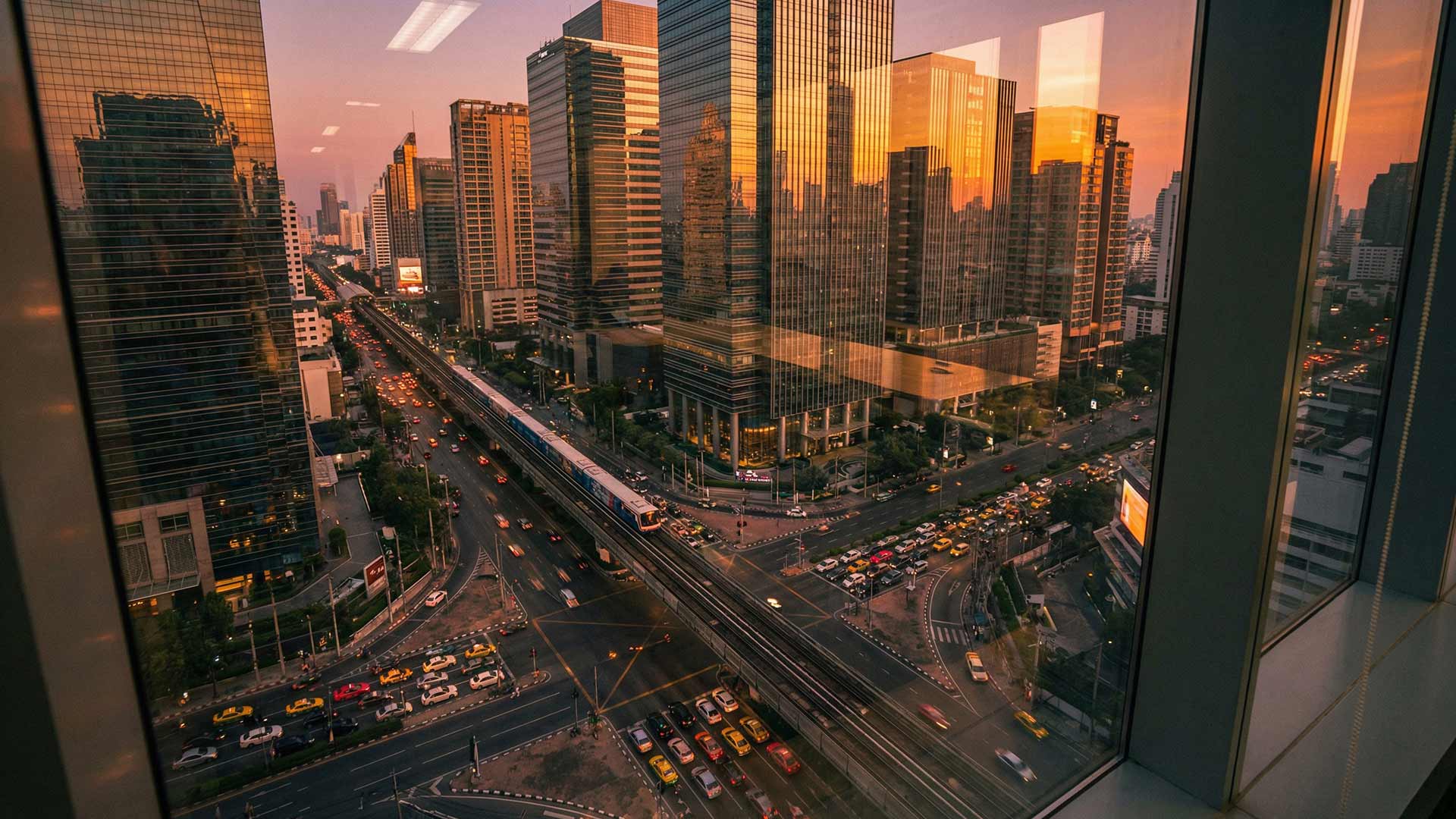 Sunset view through floor-to-ceiling windows of a high-rise office looking down on Sukhumvit Road traffic, BTS Skytrain, and glass tower reflections